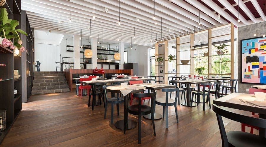 Interno moderno del ristorante Myricae a Jesolo, con arredi minimalisti in legno, tavoli apparecchiati, sedie nere e rosse, luci a sospensione e un grande quadro colorato sulla parete.